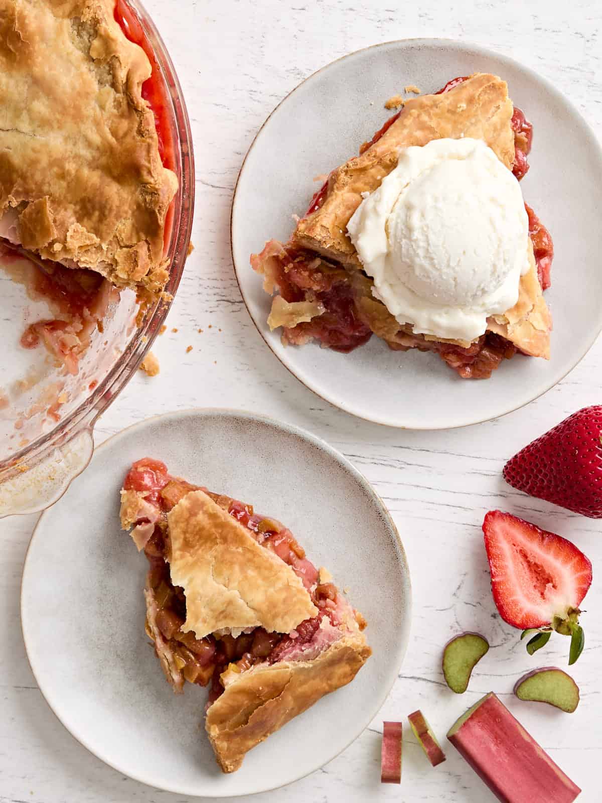 Overhead view of two slices of strawberry rhubarb pie on two plates, with one topped with ice cream.