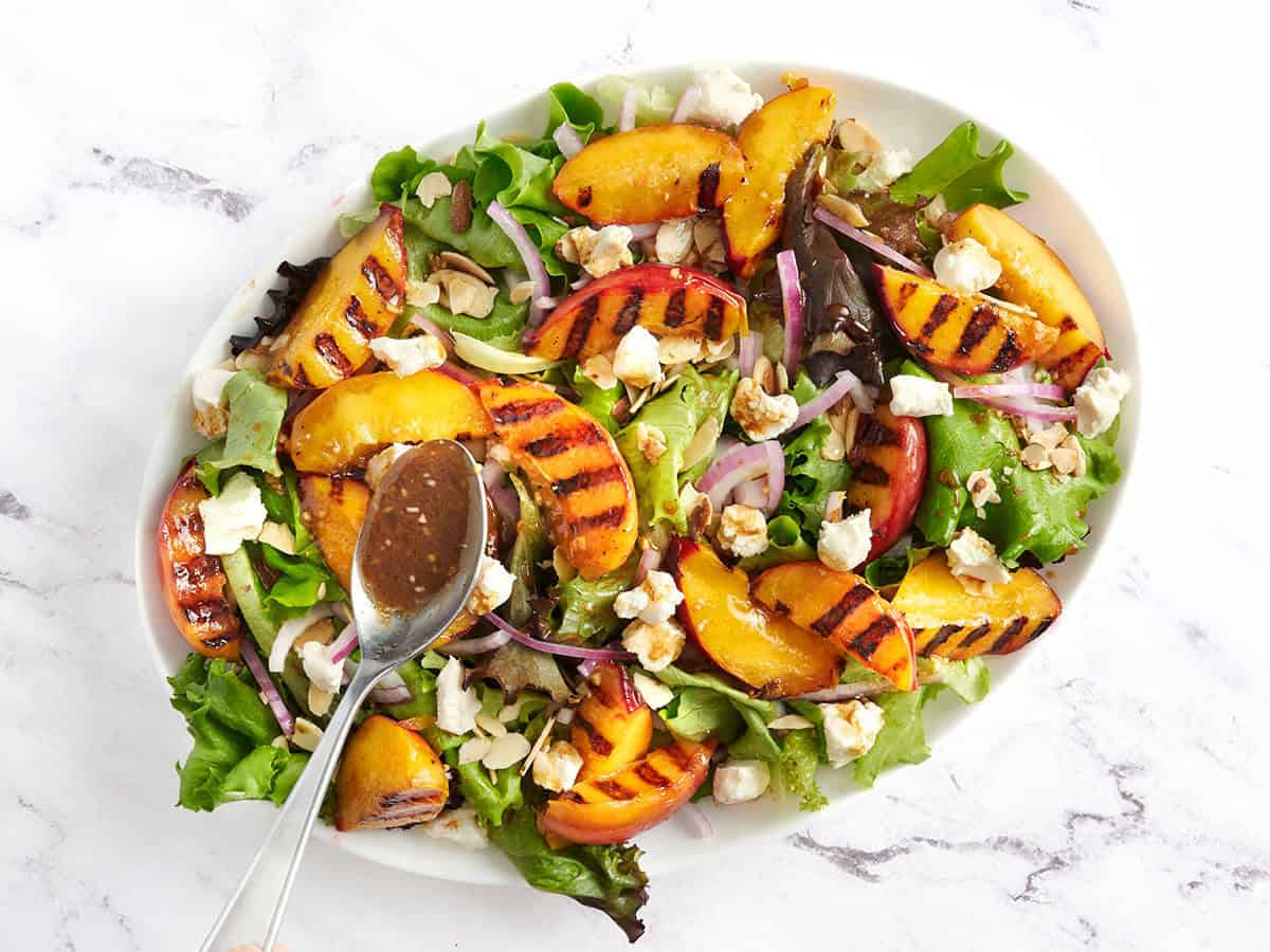 A spoon adding dressing to a platter of grilled peach salad.