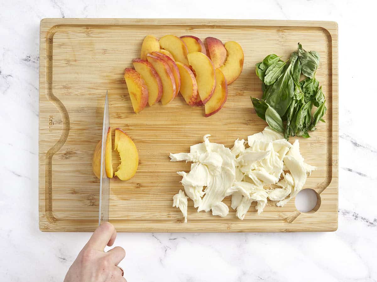 A knife slicing peaches on a wooden cutting board next to fresh mozzarella and basil leaves,