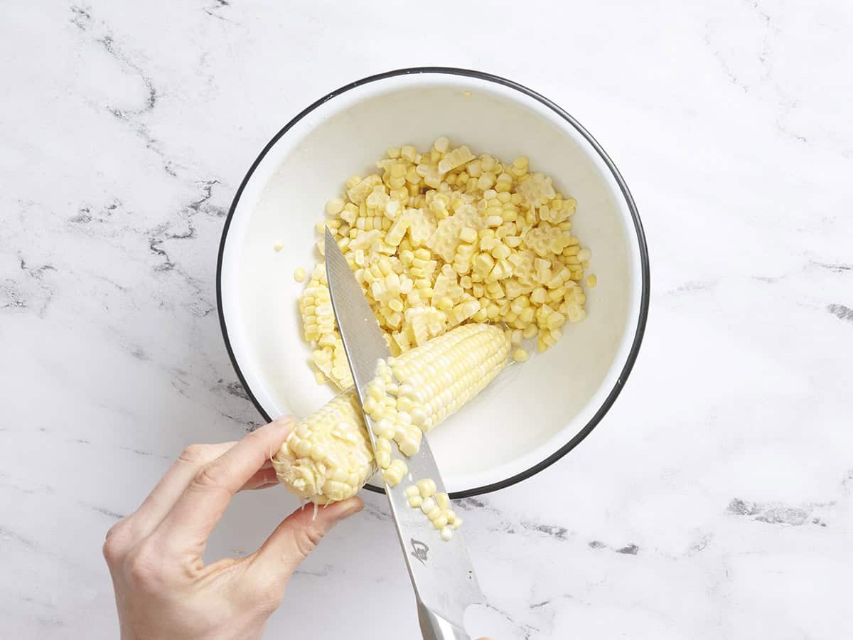 A knife shaving corn off a corn cob into a bowl.