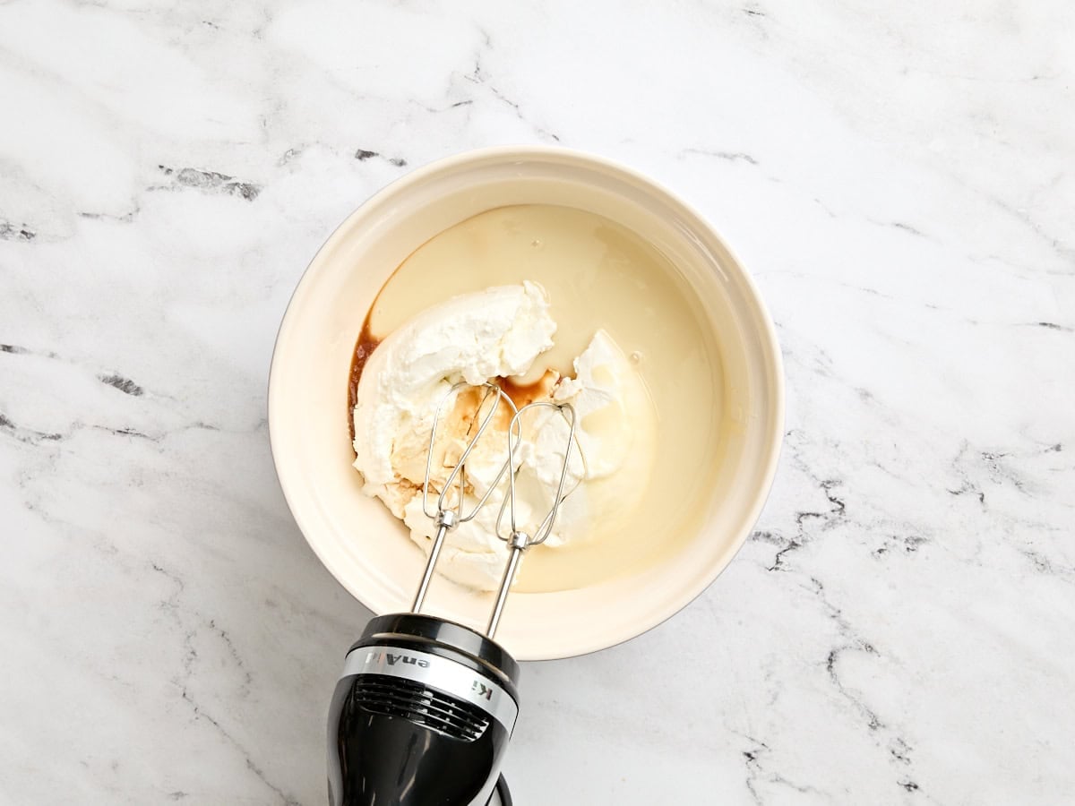 Sweetened condensed milk, greek yogurt, and vanilla extract being mixed with a hand mixer in a mixing bowl.