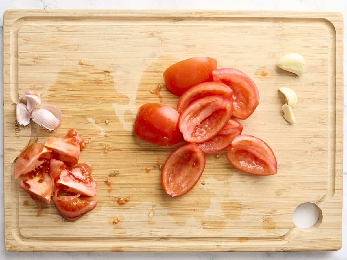 A deseeded tomato and garlic cloves on a wooden cutting board.