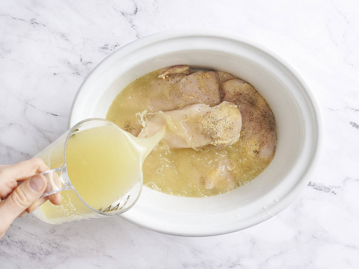 Chicken breasts in a crockpot with chicken broth being poured on top.