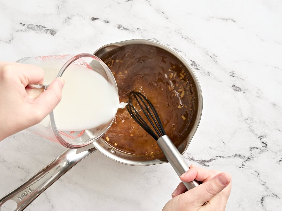 A cornstarch slurry being poured into homemade teriyaki sauce in a sauce pot.