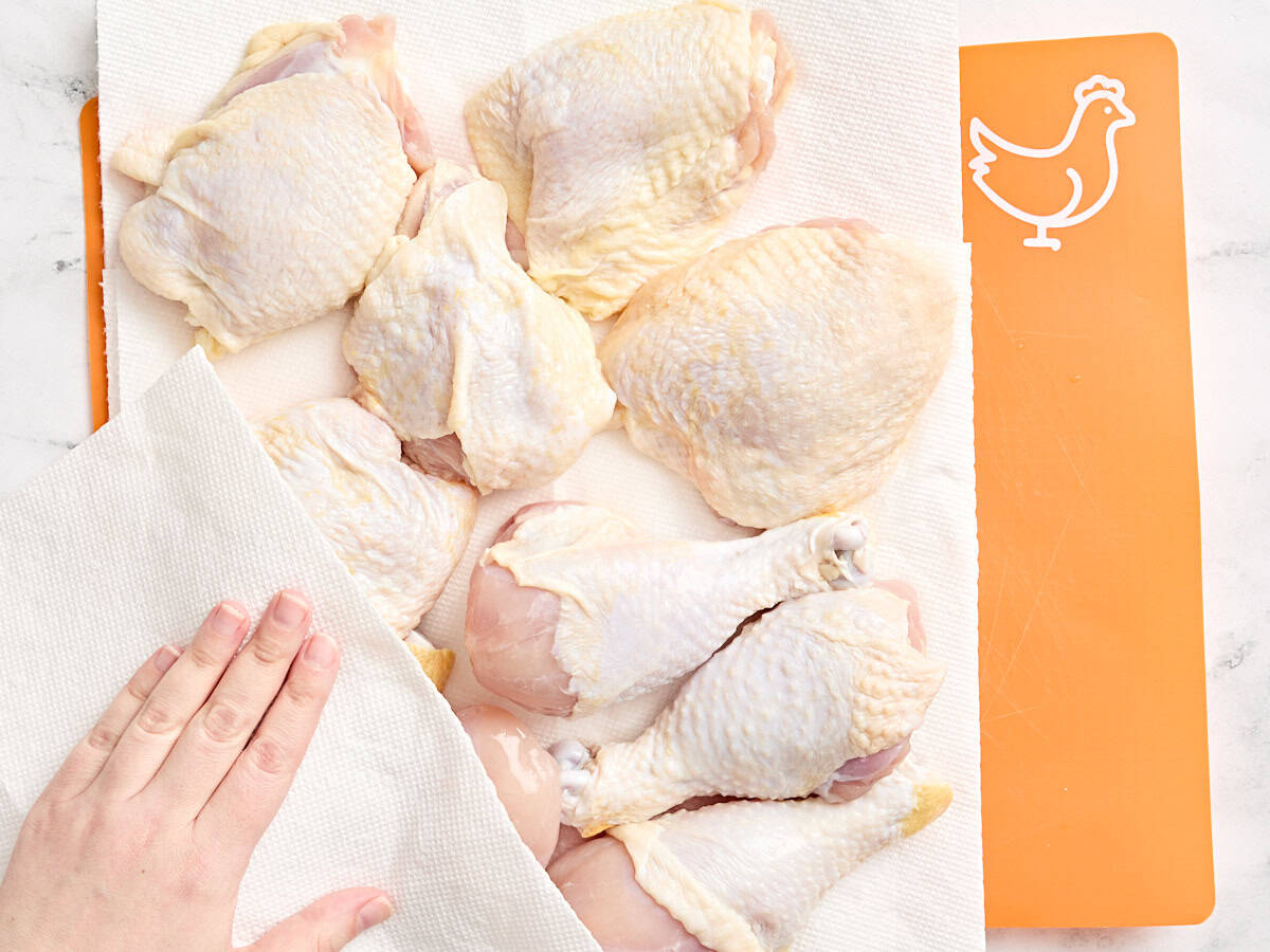 A hand using a paper towel to pat chicken drumsticks and thighs dry.