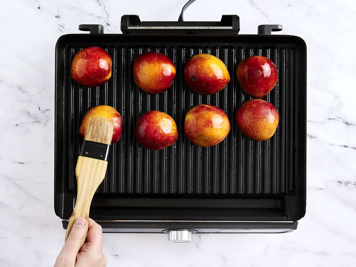 A hand brushing peach halves on the grill with oil.