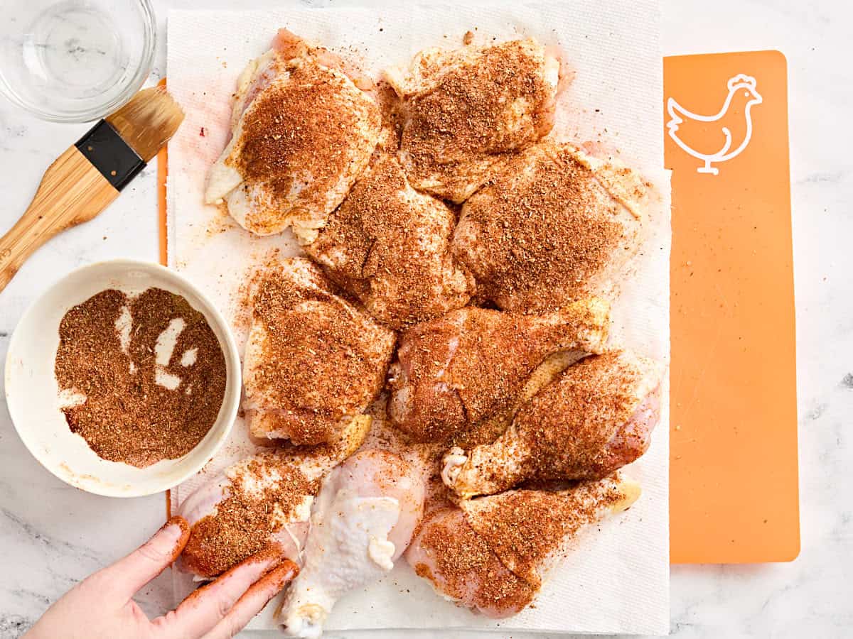 A hand rubbing a dry rub on chicken drumsticks and thighs.
