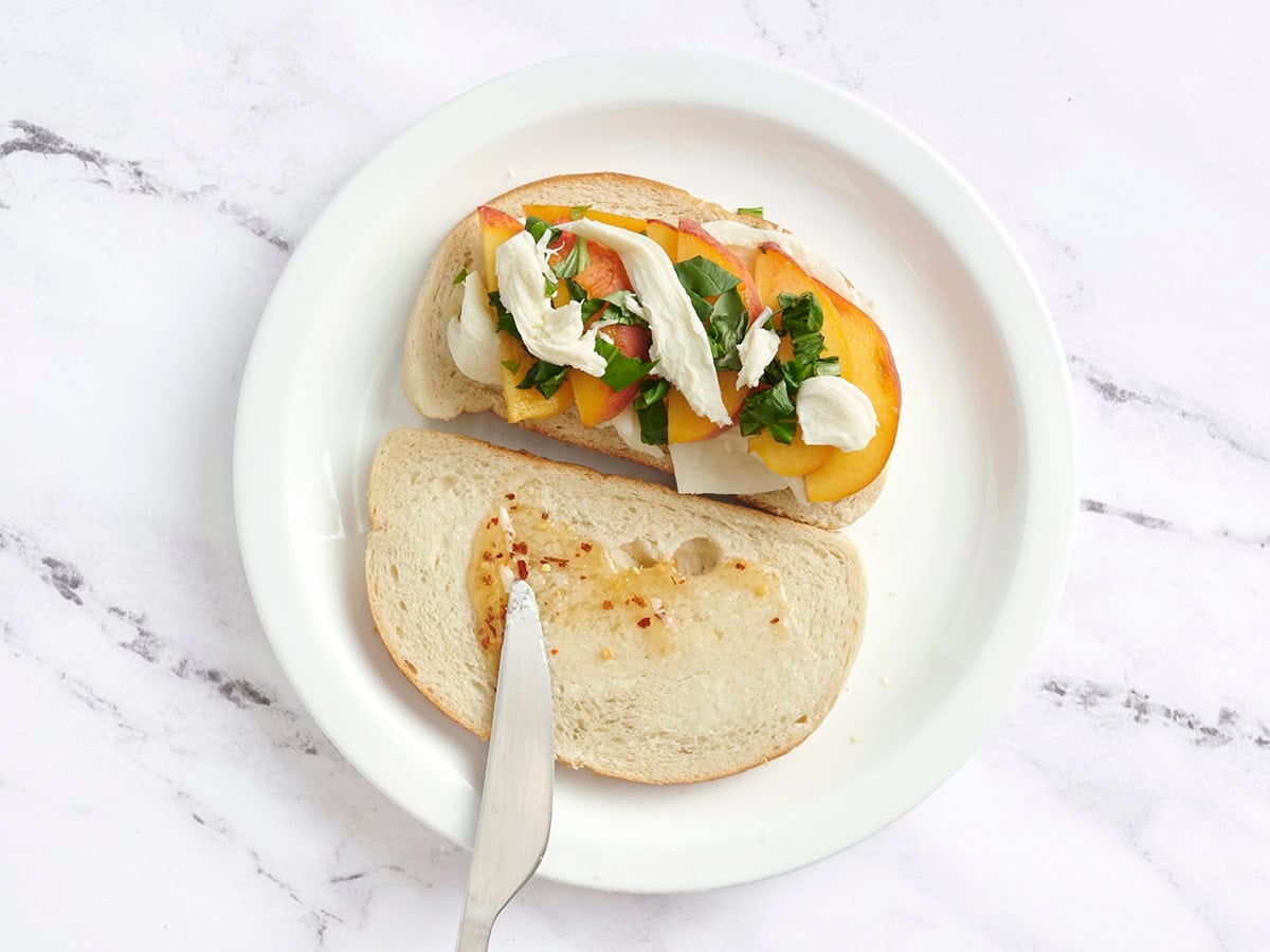 Two slices of buttered sourdough on a plate, with one topped with peaches, mozzarella and basil and the other is having honey honey spread on top.