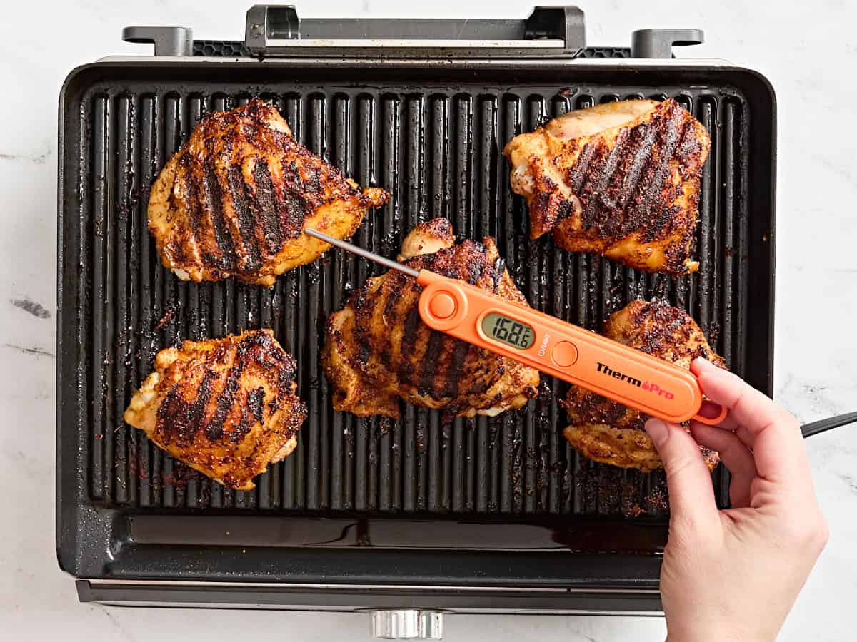 A meat thermometer checking the internal temperature of grilled chicken thighs on a grill.