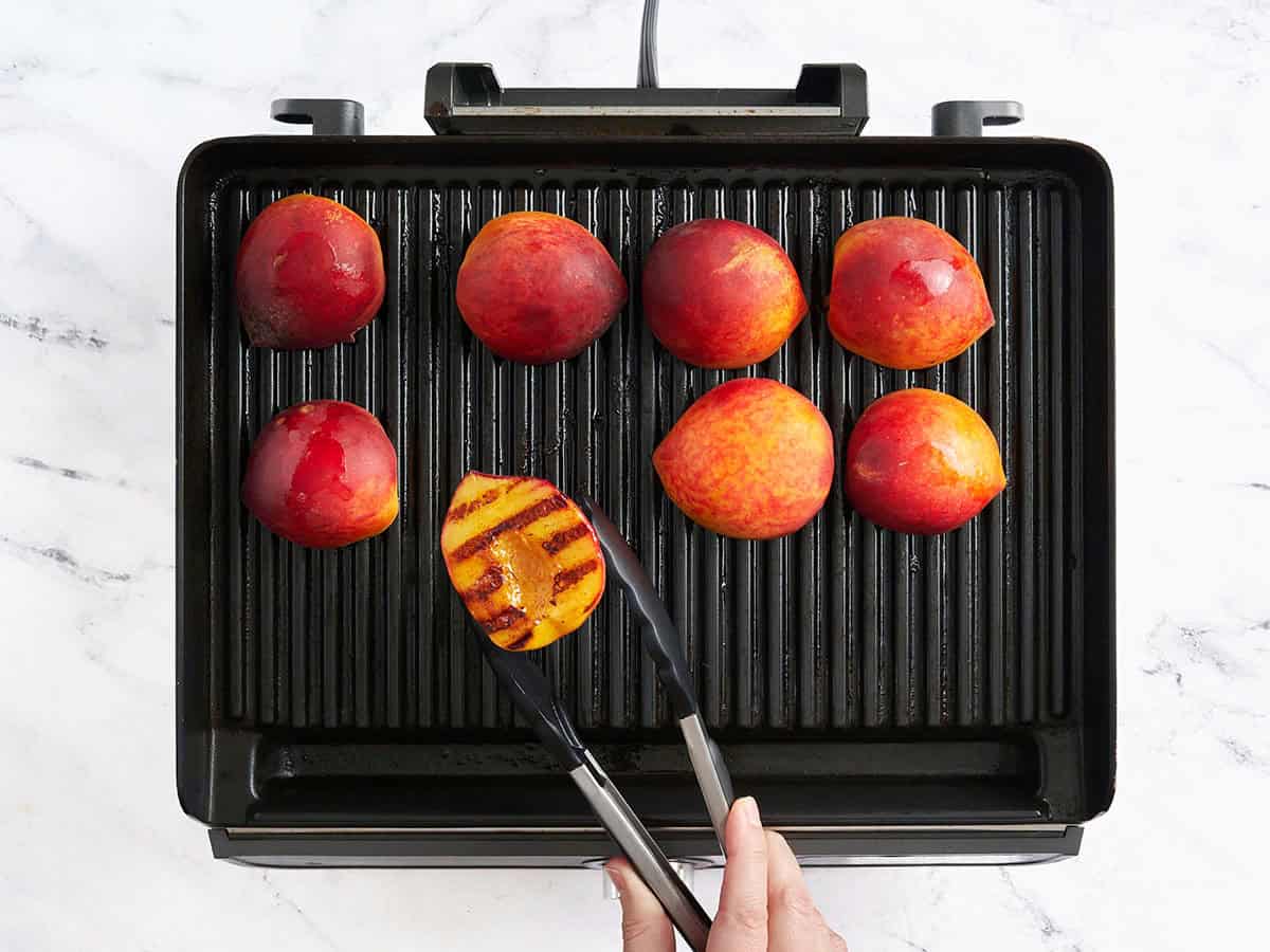 Kitchen tongs flipping grilled peaches over on a grill.
