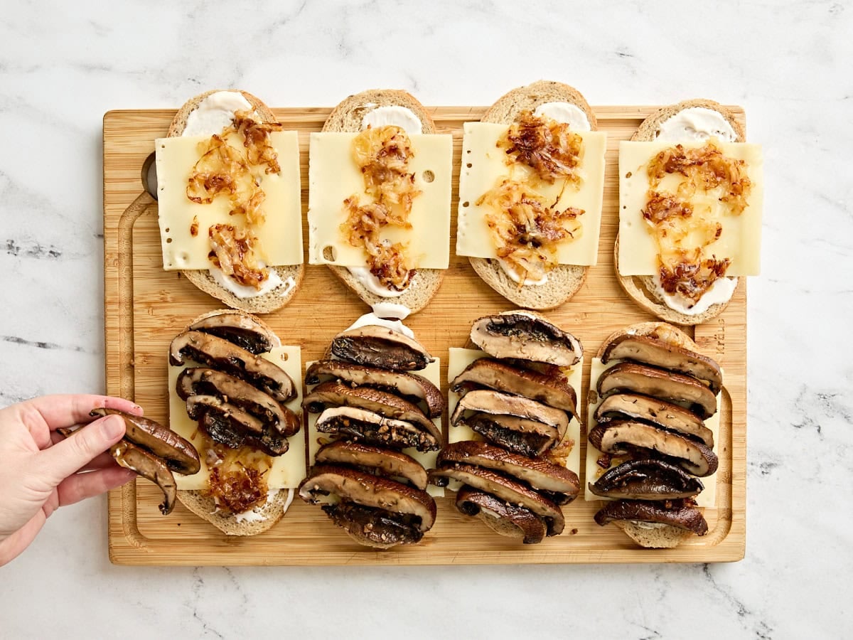 A hand adding portabella slices to a sandwich on a wooden cutting board.