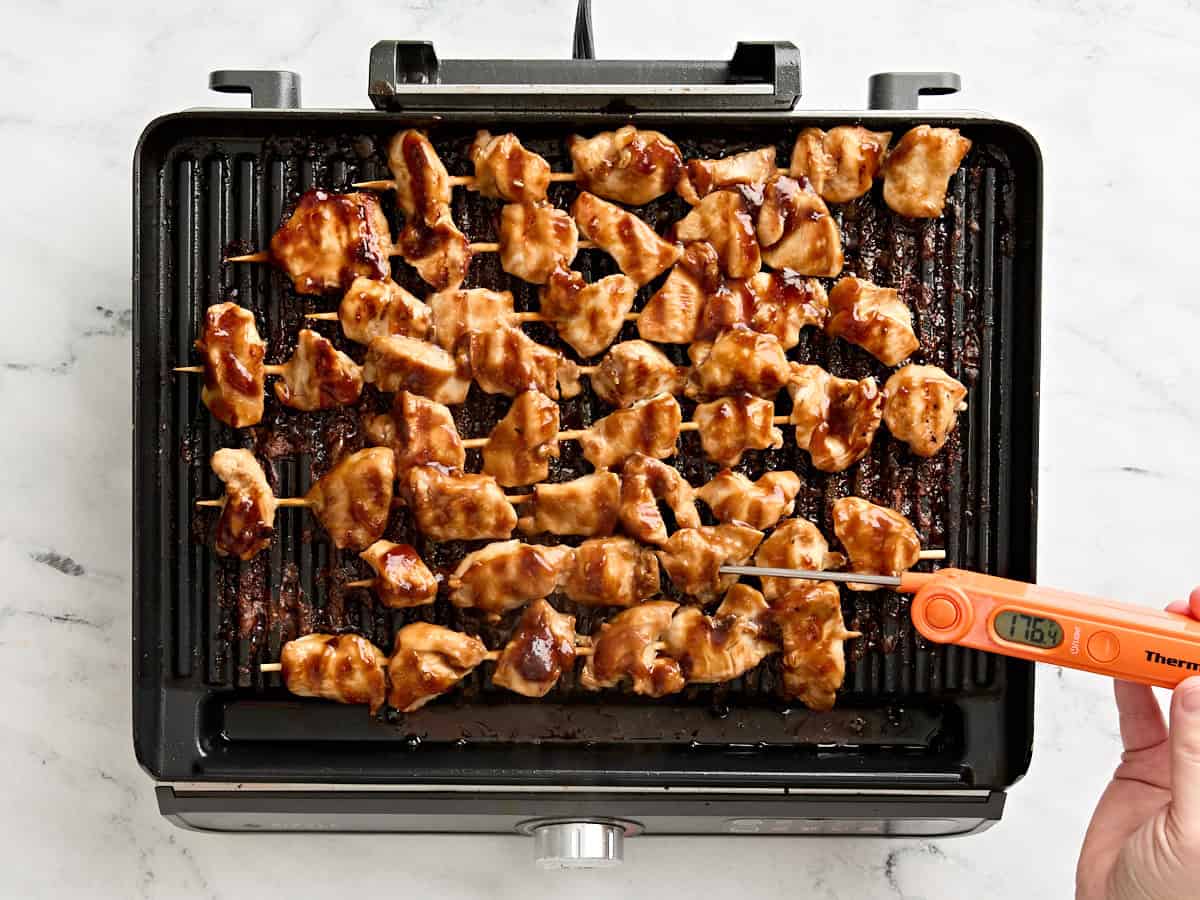 A meat thermometer checking the internal temperature of grilled chicken kebabs on a grill.