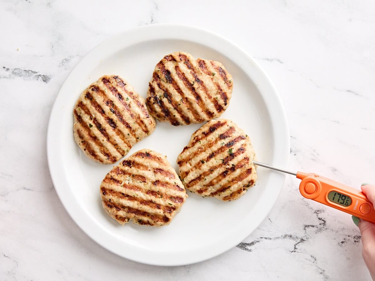 A meat thermometer checking the internal temperature of grilled chicken burgers.
