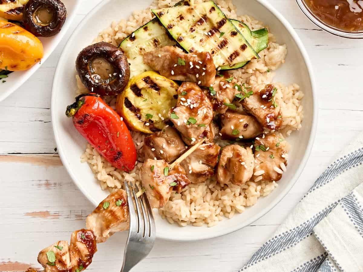 Overhead view of a plate of cooked rice with grilled vegetables. A fork is also removing the chicken from a grilled teriyaki chicken skewer onto the plate.