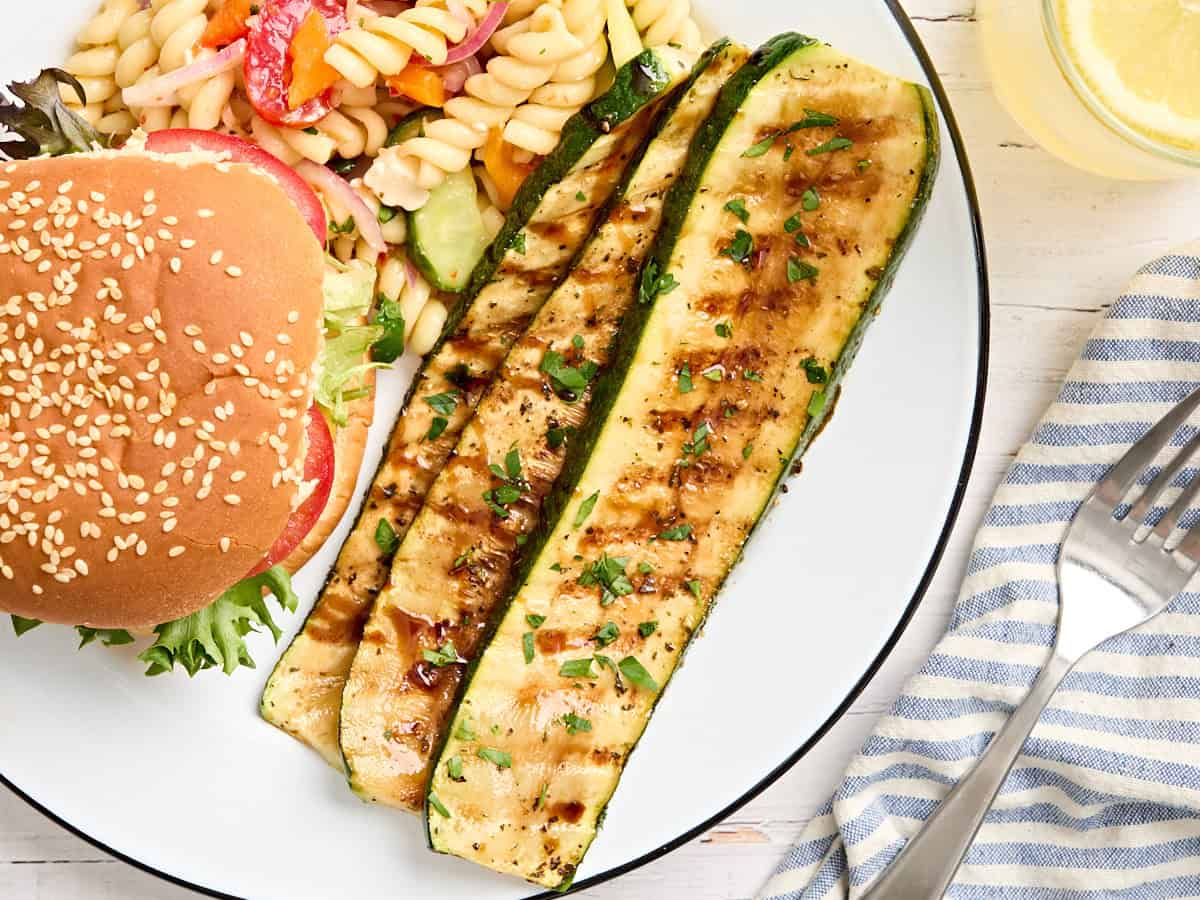 Overhead view of grilled zucchini slices on a plate with pasta salad and a burger.