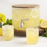 A drink dispenser with homemade lemonade and two glasses of lemonade in front.