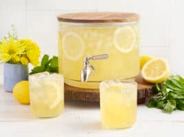 A drink dispenser with homemade lemonade and two glasses of lemonade in front.
