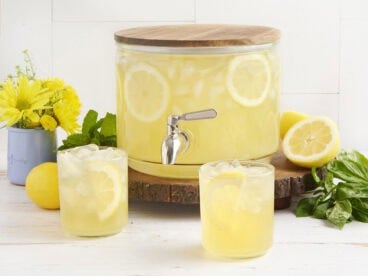 A drink dispenser with homemade lemonade and two glasses of lemonade in front.