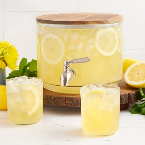 A drink dispenser with homemade lemonade and two glasses of lemonade in front.