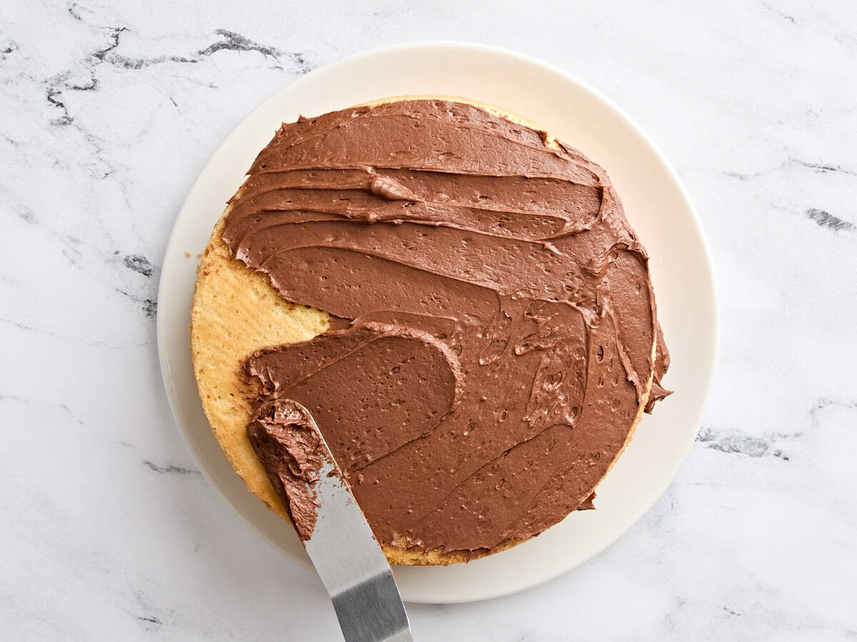 An offset spatula spreading chocolate buttercream onto a vanilla cake.