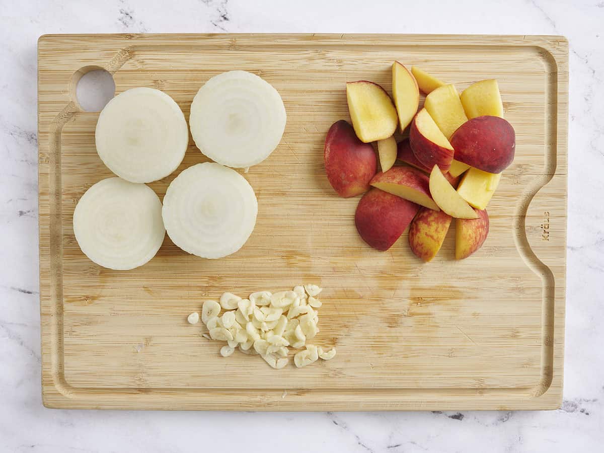 Onions sliced in half, chopped peaches, and chopped garlic on a wooden cutting board.