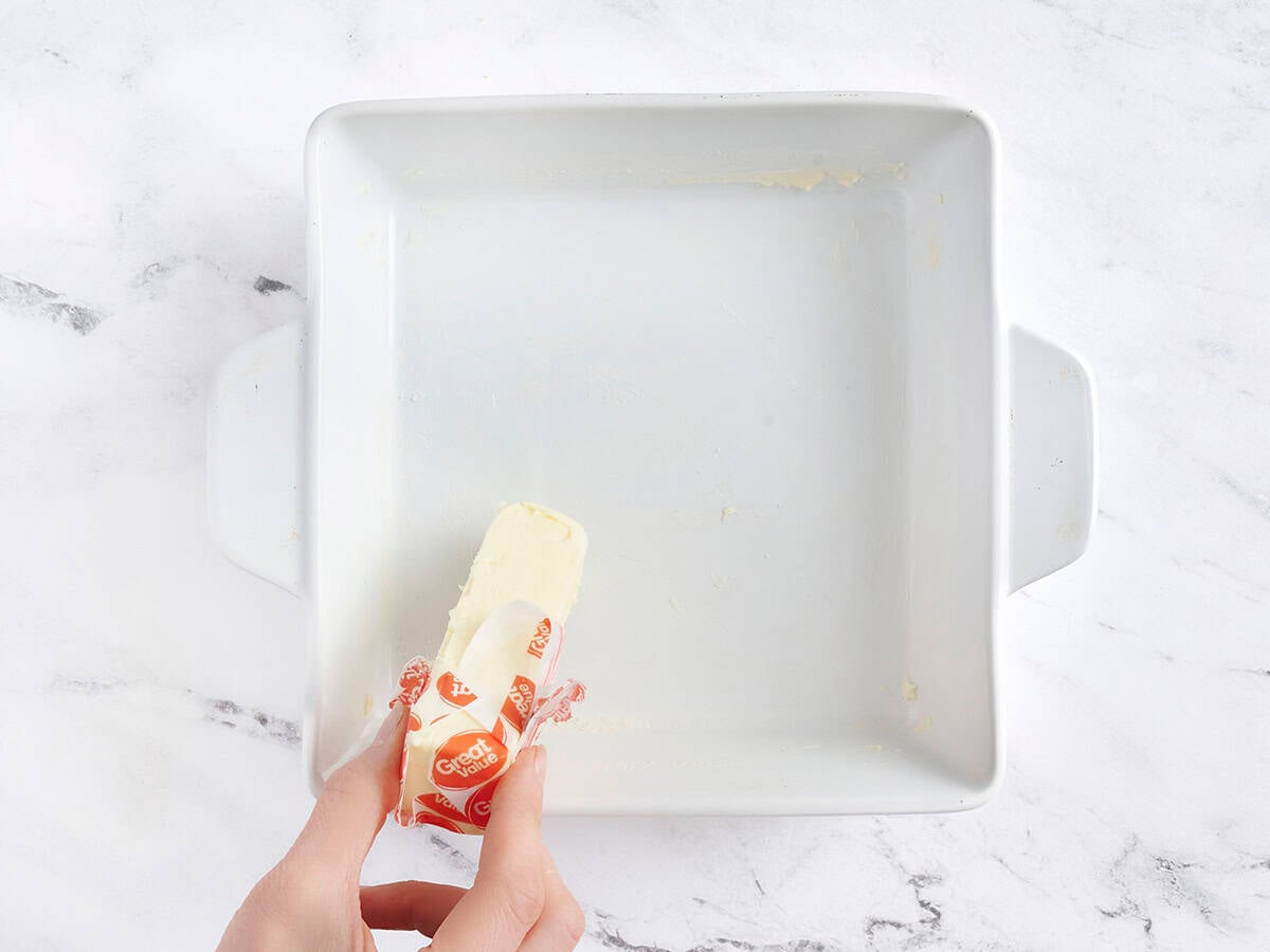 Hands using a stick of butter to grease a baking dish.