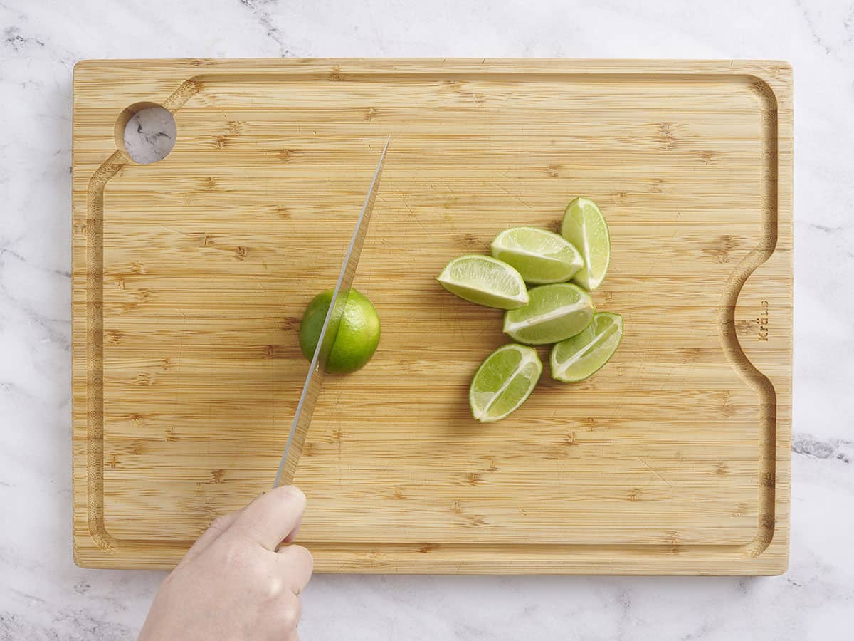 A knife slicing limes into quarters on a wooden cutting board.