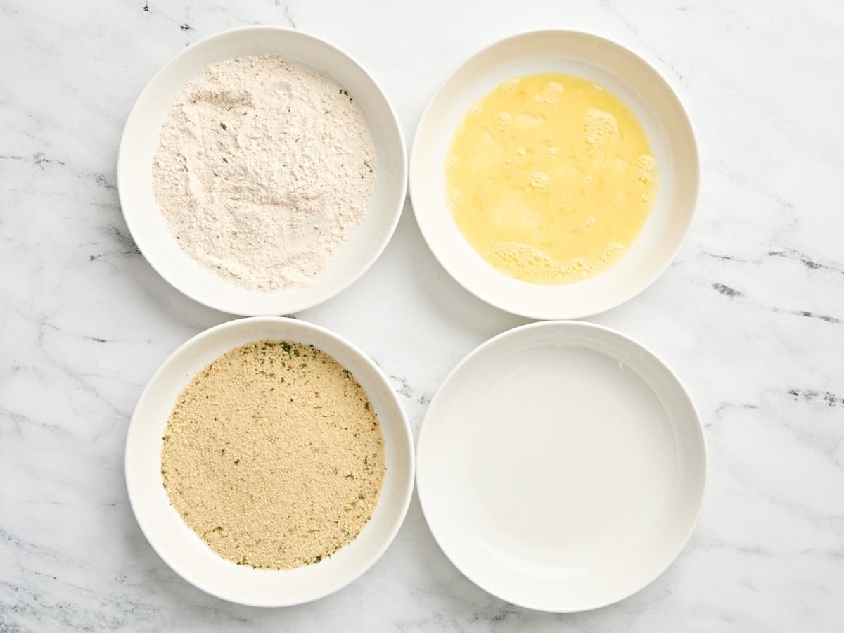 A bowl of flour, a bowl of beaten egg, a bowl of breadcrumbs, and an empty bowl.