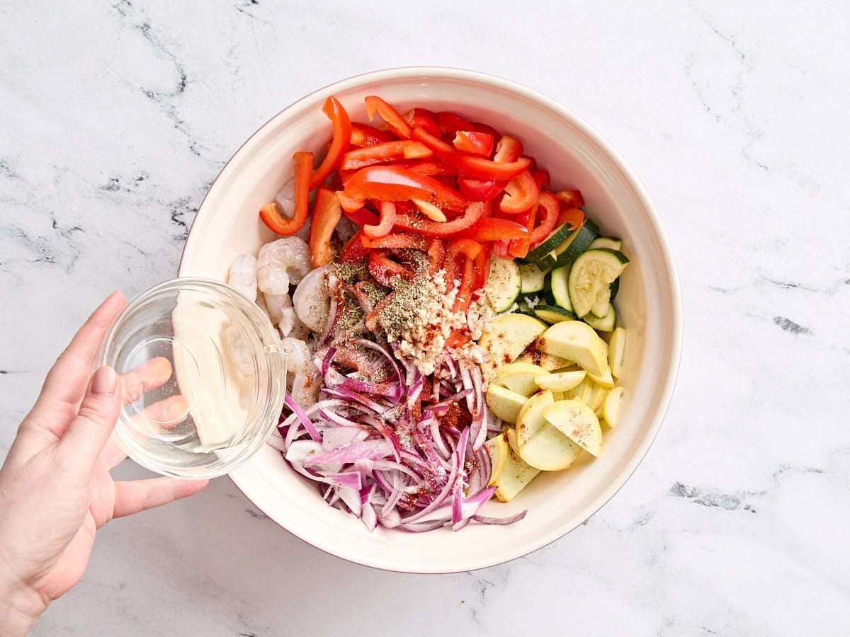 Sliced red onion, red bell pepper, yellow squash, zucchini, raw shrimp, and seasonings in a bowl, with white wine being poured on top.