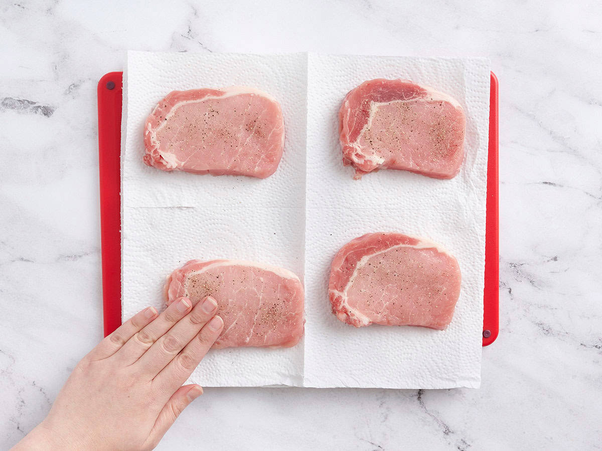 Pork chops being seasoned with salt and pepper on a paper towel.