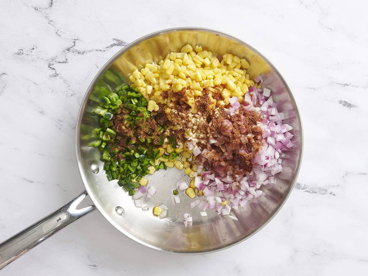Seasonings, frozen corn, diced red onion, and green pepper in a skillet.