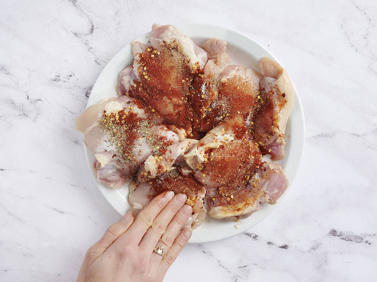 A hand rubbing seasonings onto raw chicken thighs.