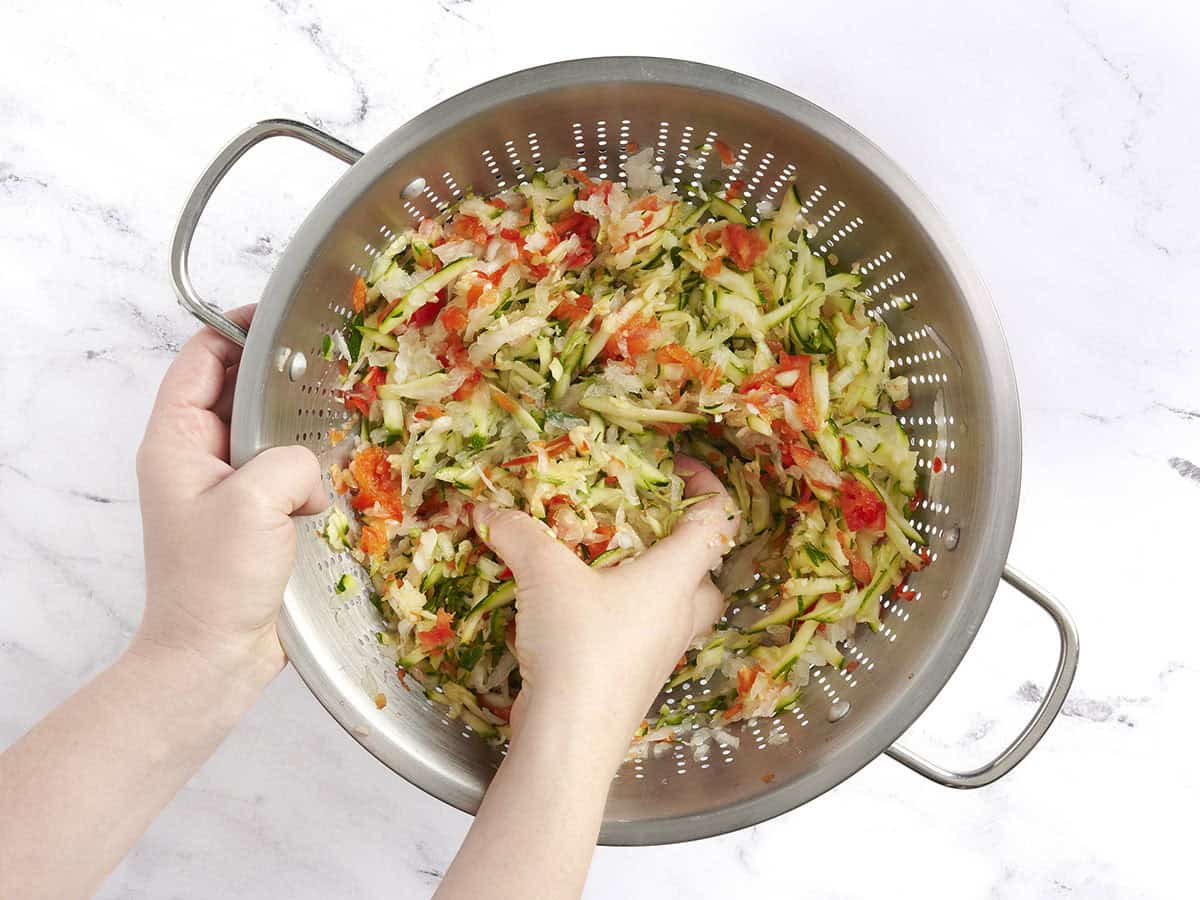 A hand massaging zucchini, onion, jalapeno, and red bell pepper in a colander.
