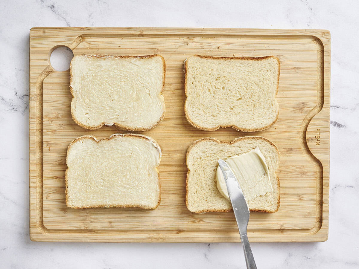 Overhead view of four slices of bread on a cutting board, with one being spread with mayo.