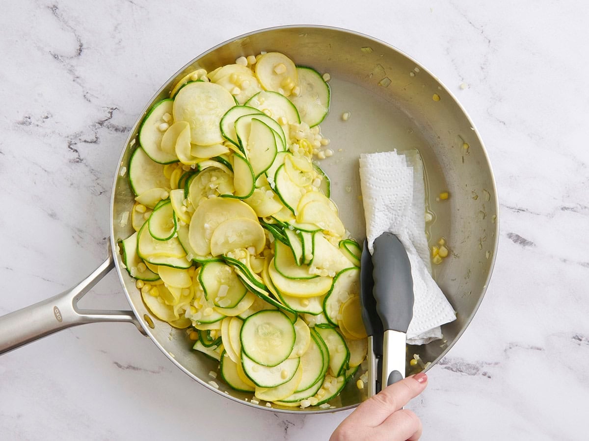 Kitchen tongs soaking up the liquid from sauteed zucchini and yellow squash in a skillet.