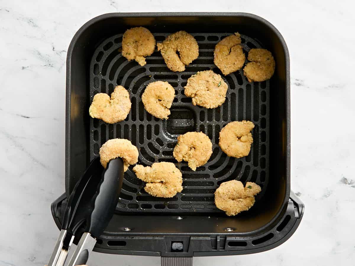Kitchen tongs flipping over air fried fried shrimp in an air fryer basket.