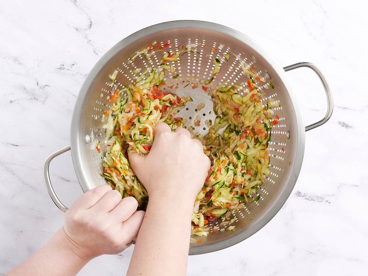 A hand squeezing out the liquid from zucchini, jalapeno, and onion in a colander.