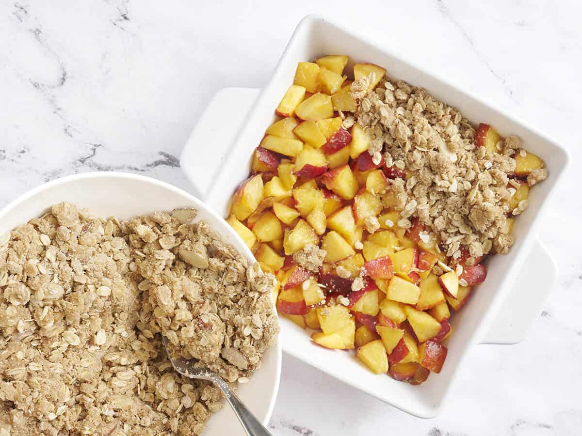Crumble topping being added to a peach filling in a baking dish.