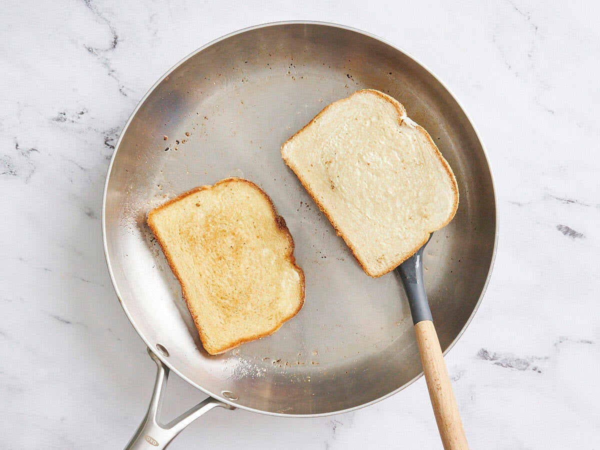Slices of bread toasting in a skillet with a spatula flipping one.