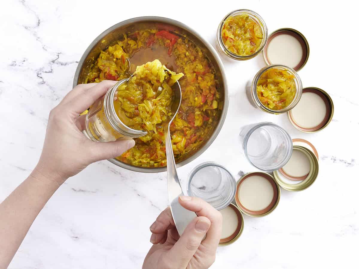 A hand spooning homemade zucchini relish into canning jars.