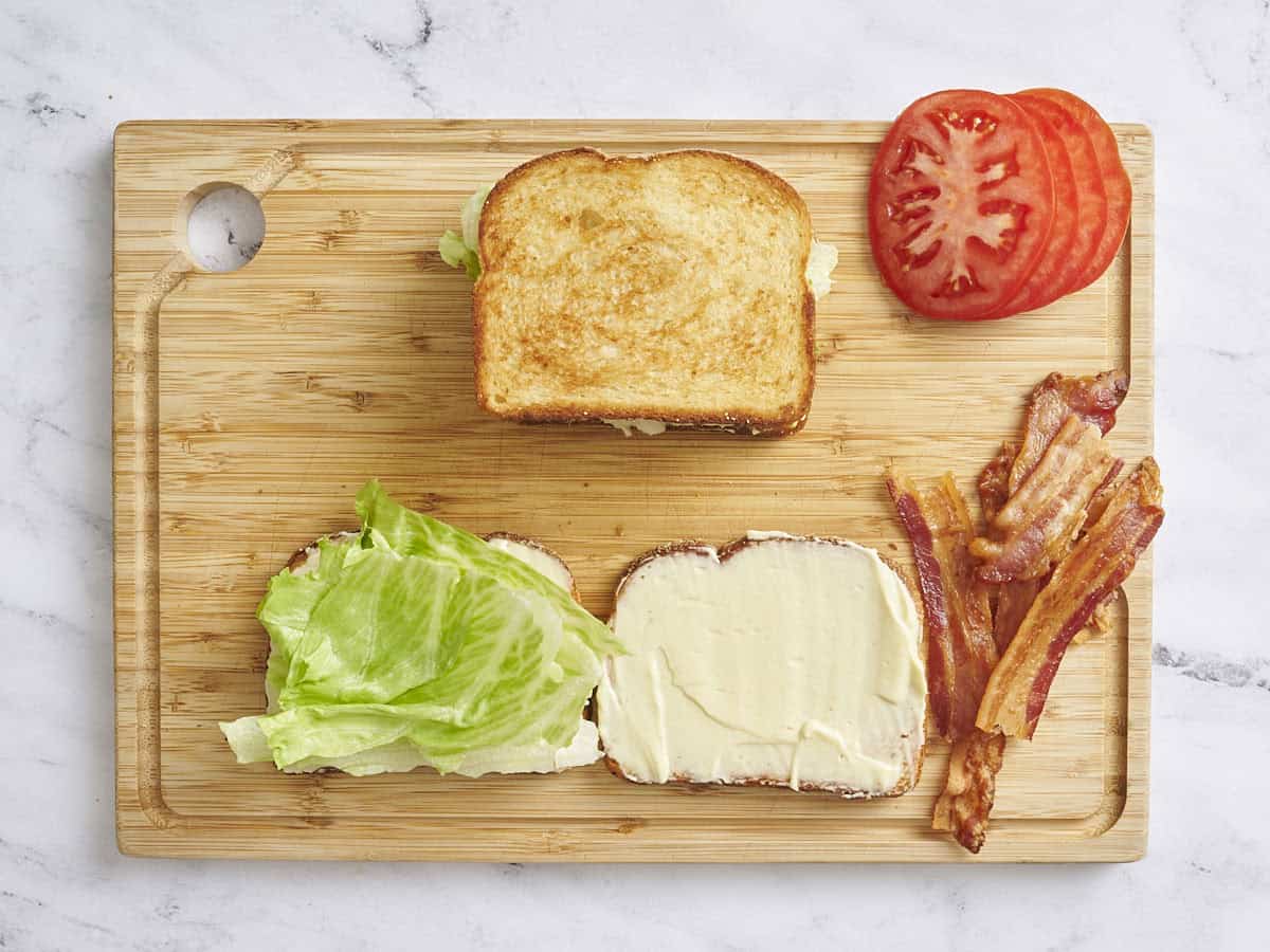 Overhead view of one assembled BLT sandwich on a wooden cutting board, with sliced tomato, cooked bacon and two slices of bread open next to it with one slice topped with lettuce.