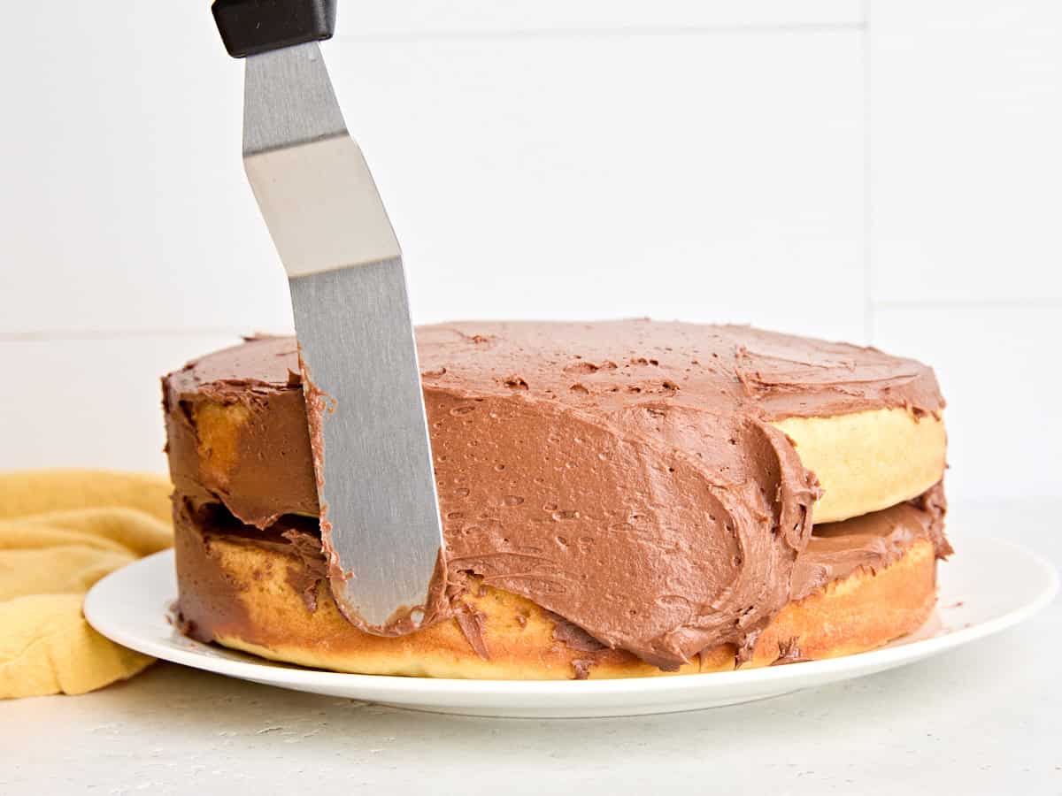 Side view of a vanilla cake being spread with chocolate buttercream frosting.