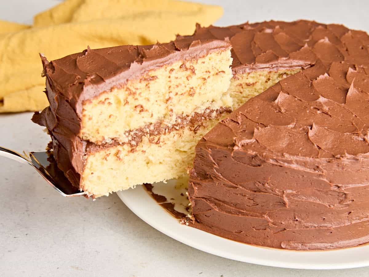 A homemade vanilla cake with chocolate frosting, with a slice being taken.