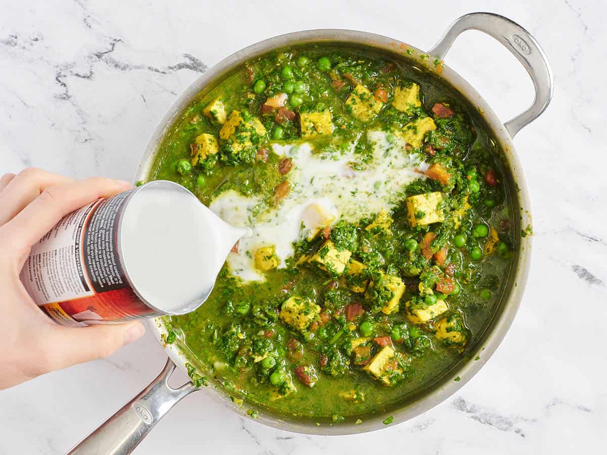 Coconut milk being poured into a skillet of tofu, spinach and green pea curry.