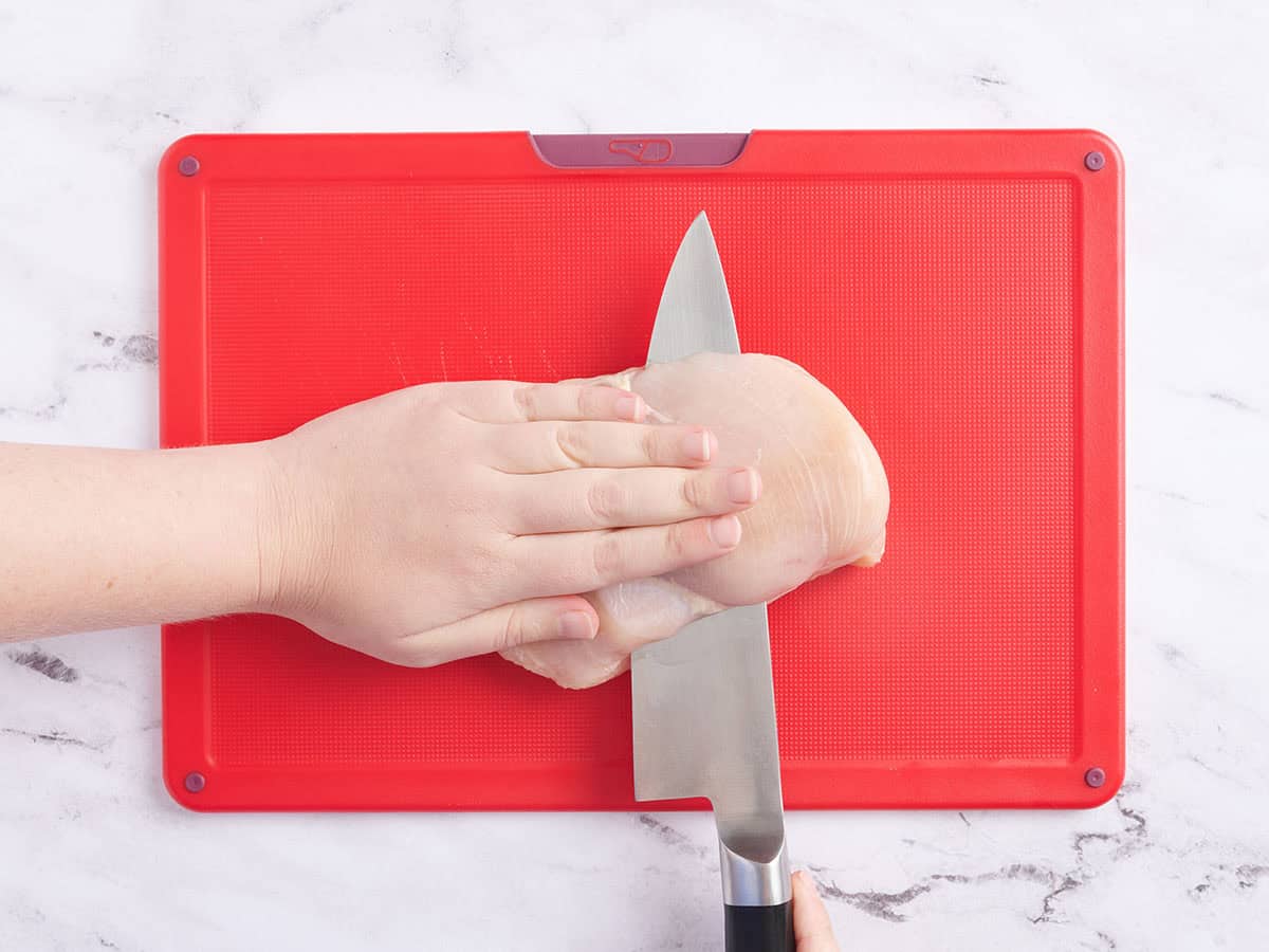 A hand using a knife to slice a chicken breast in half.
