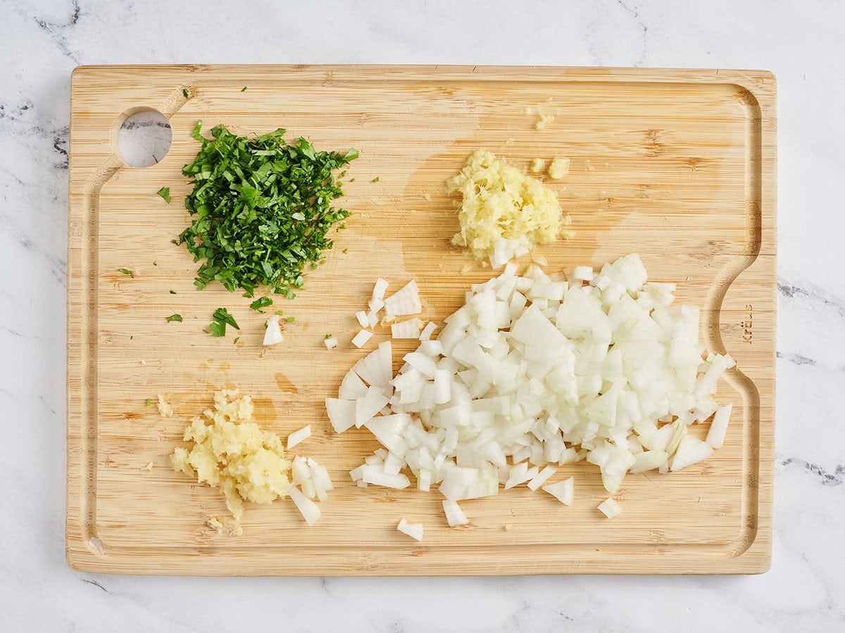 Onion, garlic, ginger, and parsley minced on a wooden cutting board.