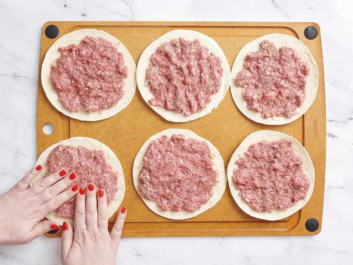 Hands pressing ground beef into flour tortillas.