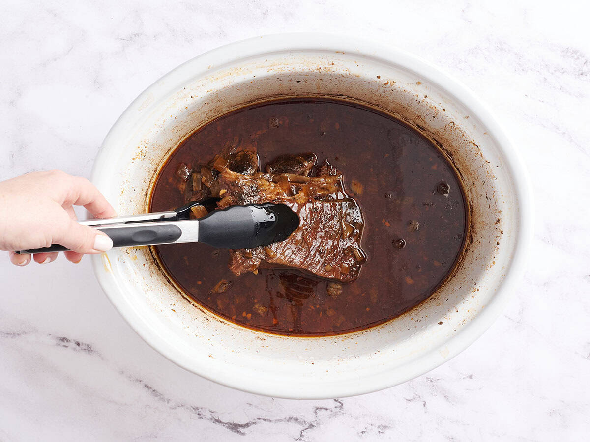 A chuck roast in a crockpot with kitchen tongs lifting it up slightly.