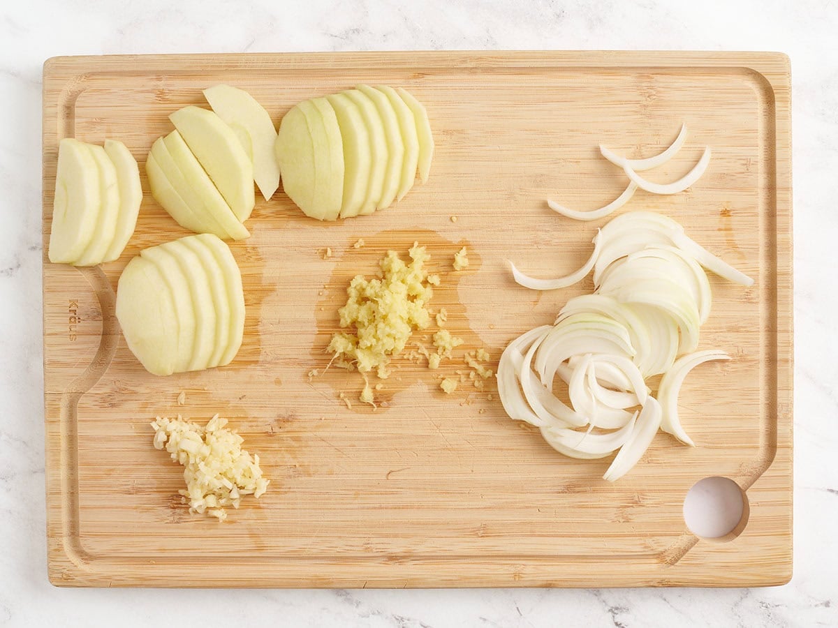 A peeled and sliced green apple, grated ginger, minced garlic, and sliced onion on a wooden chopping board.