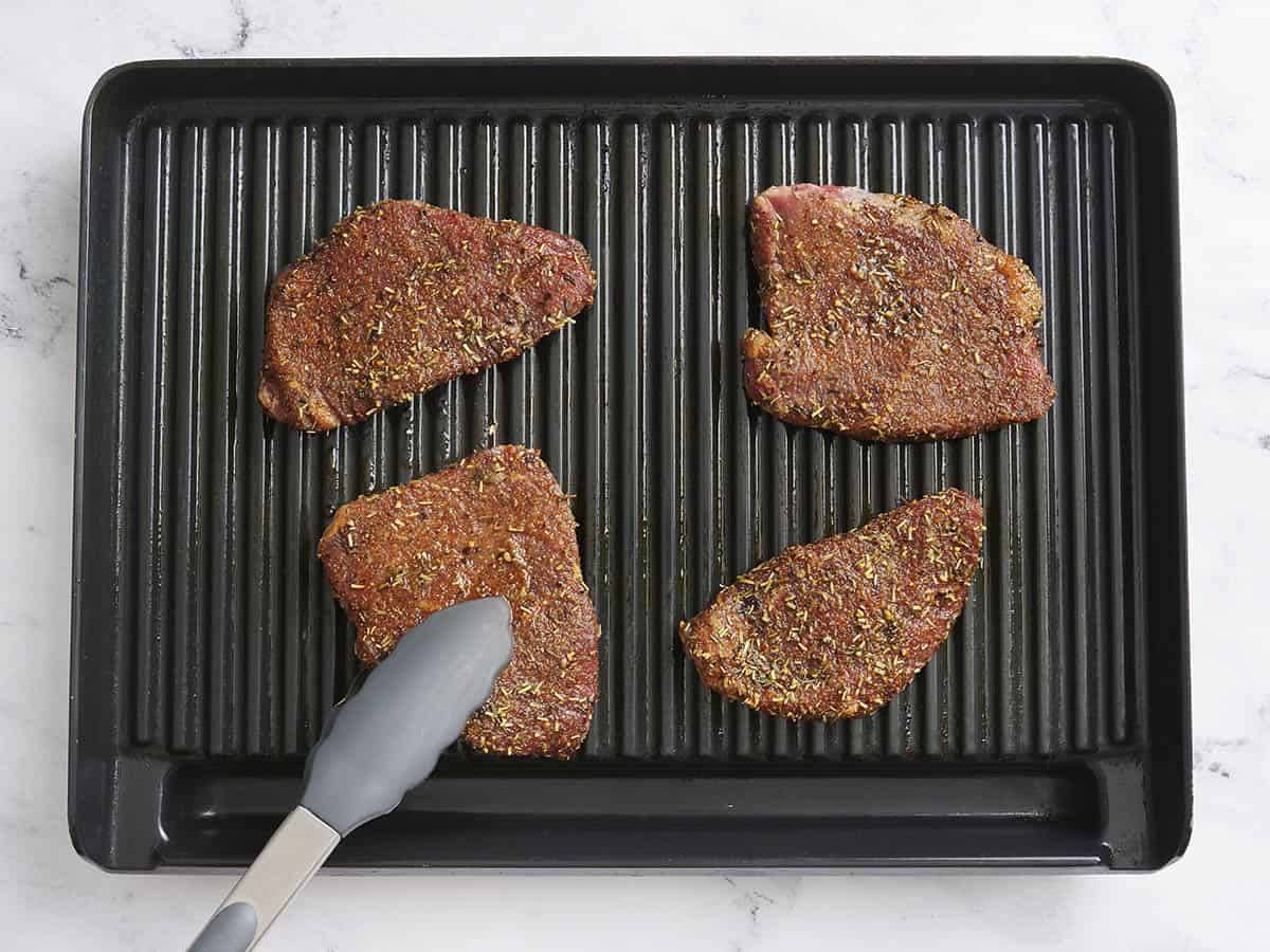 Kitchen tongs placing a sirloin tender steak on a grill.